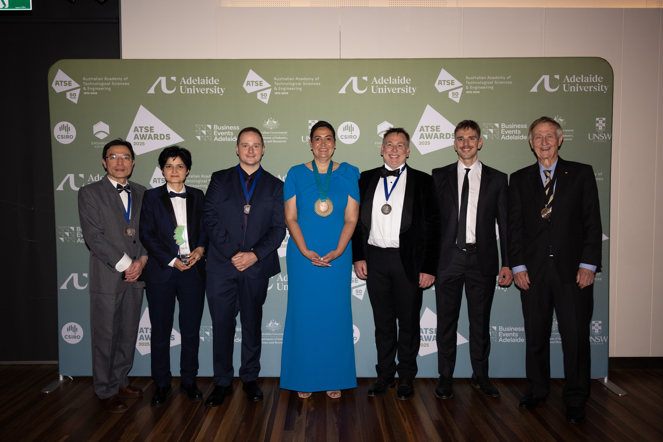 A group of 7 people, 5 men and 2 women, standing in front of a big banner featuring many logos. Most of the people are wearing medals or standing with a trophy.